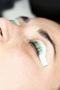 A young woman lies with closed eyes as a beautician applies eyelash extensions, exuding serenity and enjoyment. The scene is well-lit with soft, natural colors, creating a tranquil atmosphere.