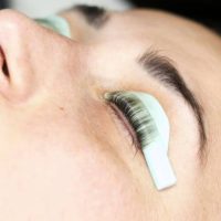 A young woman lies with closed eyes as a beautician applies eyelash extensions, exuding serenity and enjoyment. The scene is well-lit with soft, natural colors, creating a tranquil atmosphere.